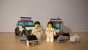 Zwei Fahrzeuge, mit einem Sanitäter und einer Ärztin. Die beiden stehen vor ihren Einsatzwagen. Vor ihnen liegt ihre Ausrüstung